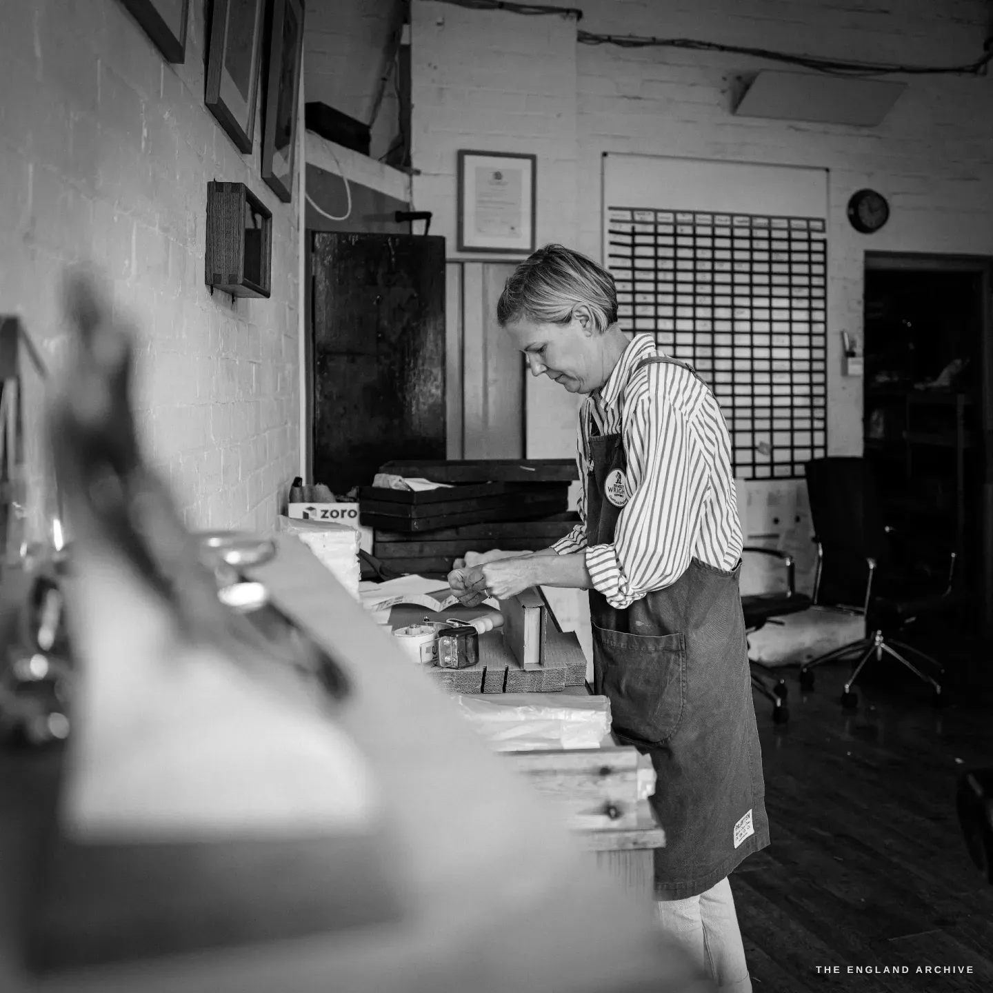 A colleague in a striped shirt and work apron bent over the packing bench, wrapping finished scissors, framed certificates on the brick wall behind.