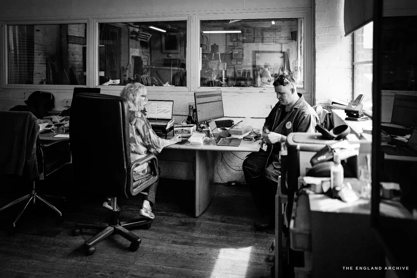 The office from another angle: the two staff at their desks and a maker sat at the end of the table, the workshop through the internal window.