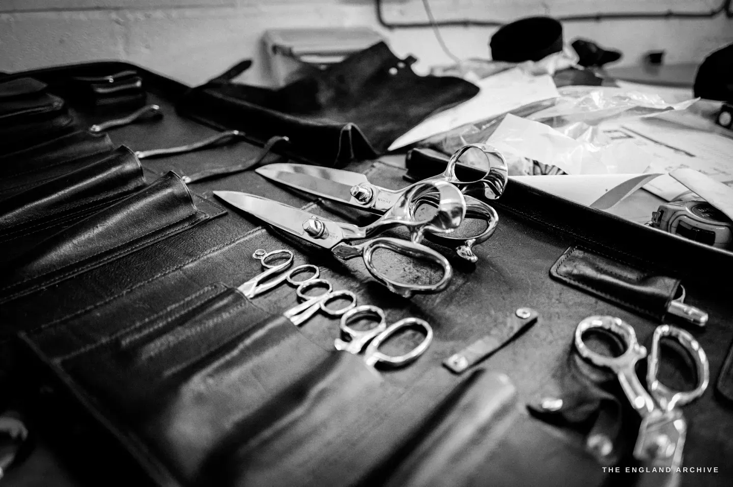 A dark leather tool roll opened out on a bench with finished scissors and embroidery bows laid across it, a tape measure to one side.
