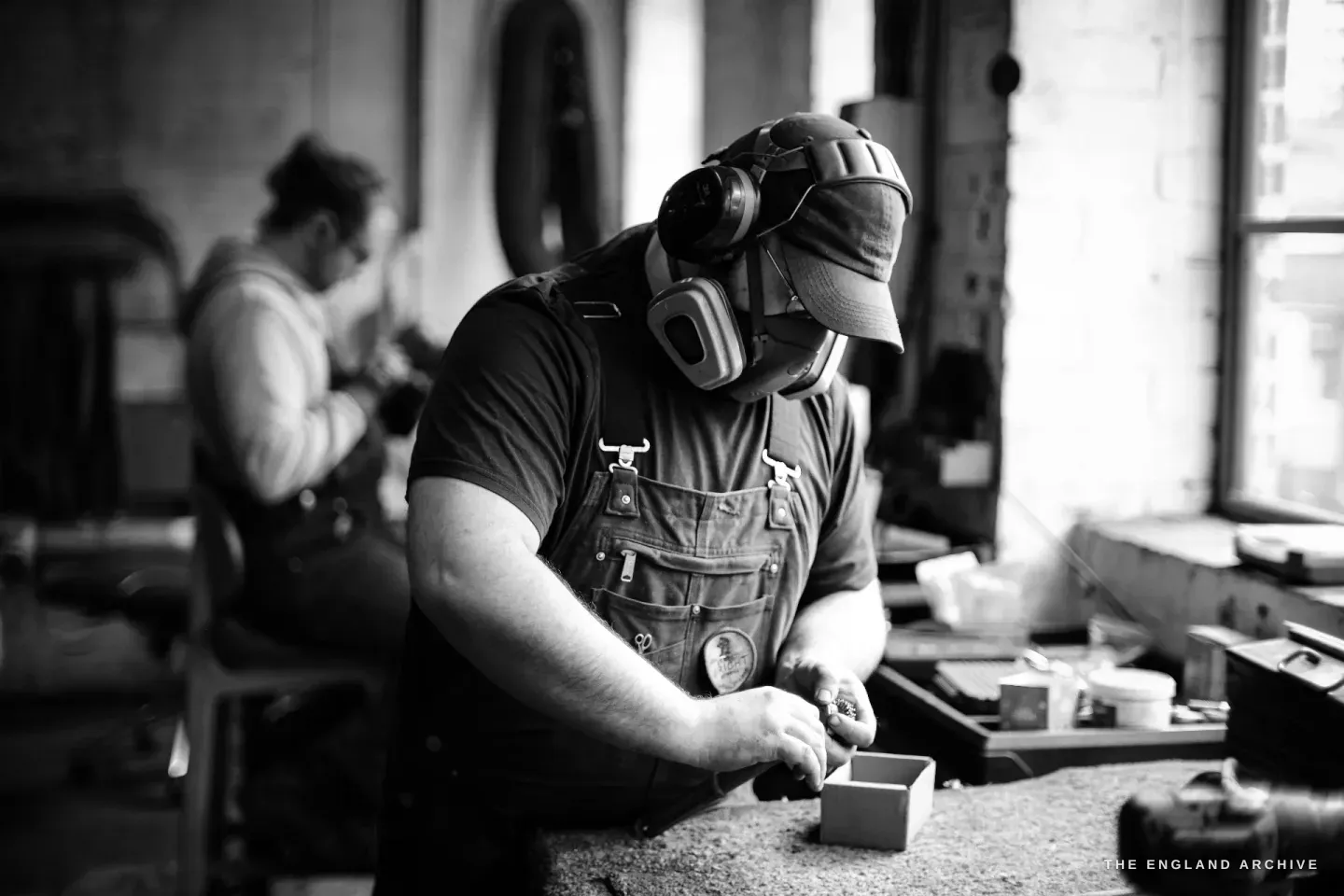 Evan James bent over a bench in a cap and ear defenders, working a small steel part with both hands, another maker at a station behind him.