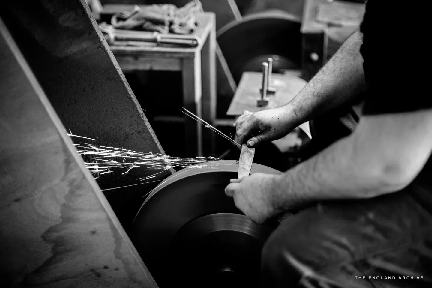 A blade held flat against a spinning grinding wheel, a bright fan of sparks streaming off to the left, two hands steadying the steel.