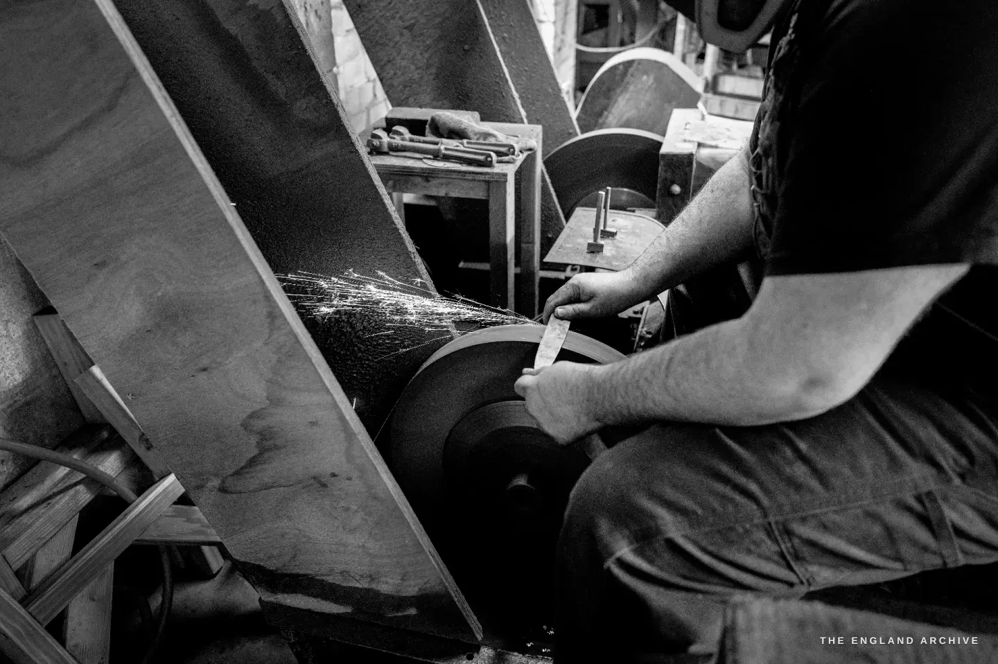 Seen from behind, Evan seated astride the grinding trough holding a blade to the wheel, sparks streaming across the dark guard.