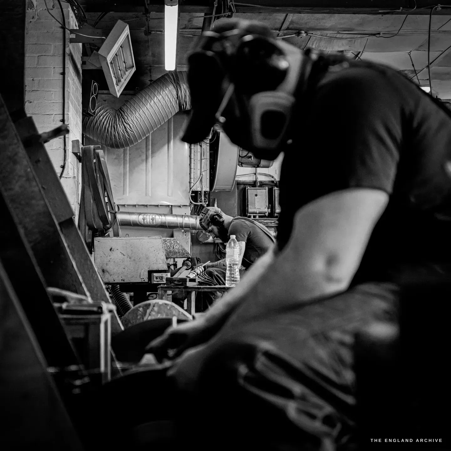 Evan in ear defenders at his grinding wheel in the foreground, the line of grinding stations and overhead extraction ducting receding behind him.