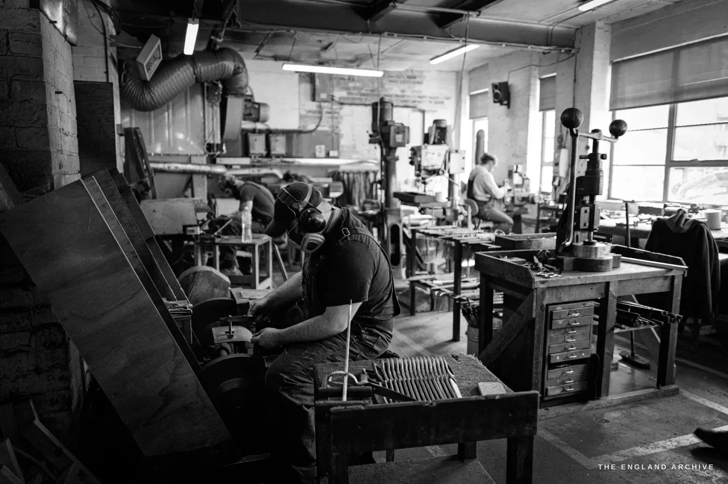 A wide view of the Ernest Wright grinding shop: Evan at a wheel mid-floor, racks of blades on the bench, other makers and a window beyond.