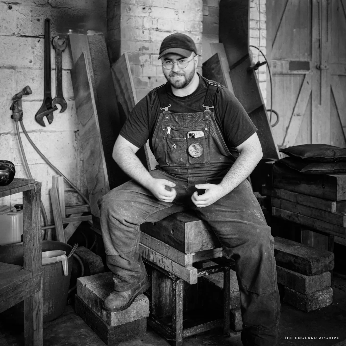 Evan James seated astride the wooden grinding horse in a cap and glasses and dungarees, looking to camera, a large wrench hanging on the wall behind him.