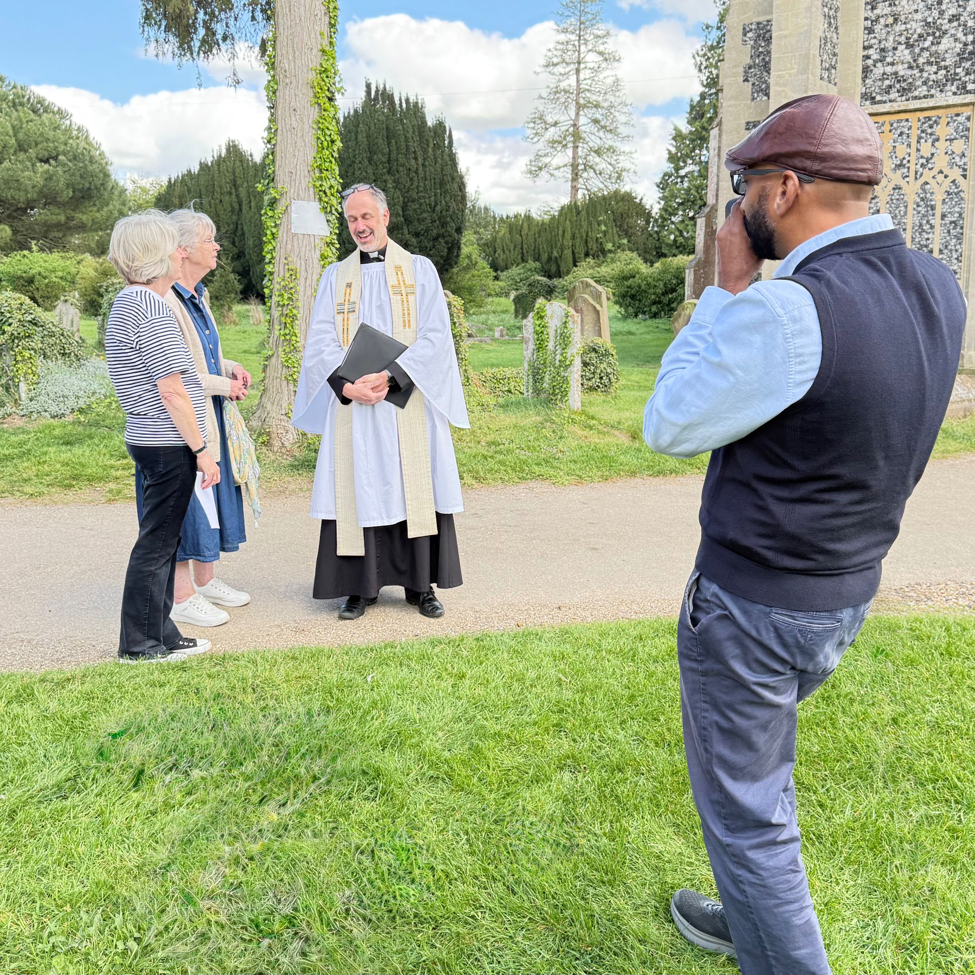 The Reverend Matthew Lawson, Rector of Holy Trinity, Long Melford, in white surplice and stole, stands in the churchyard talking with Julie Thomson and Melonie Clubb; Mash is visible on the right with a camera raised, photographing the moment.