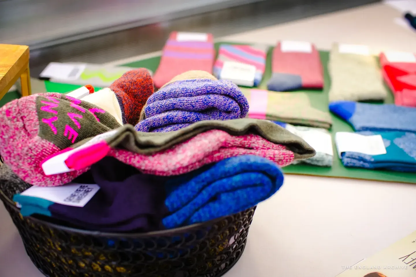 A wire basket heaped with rolled socks in pink, purple, blue and green, more pairs laid out on a table behind.
