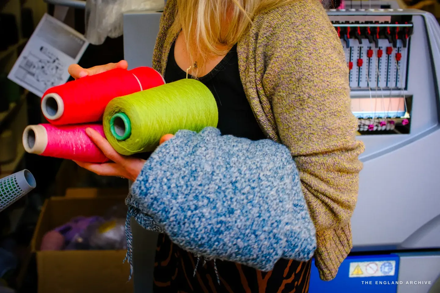 Genevieve carrying an armful of yarn cones in red, lime green and pink, a soft blue scarf folded over her arm, beside the knitting machine.