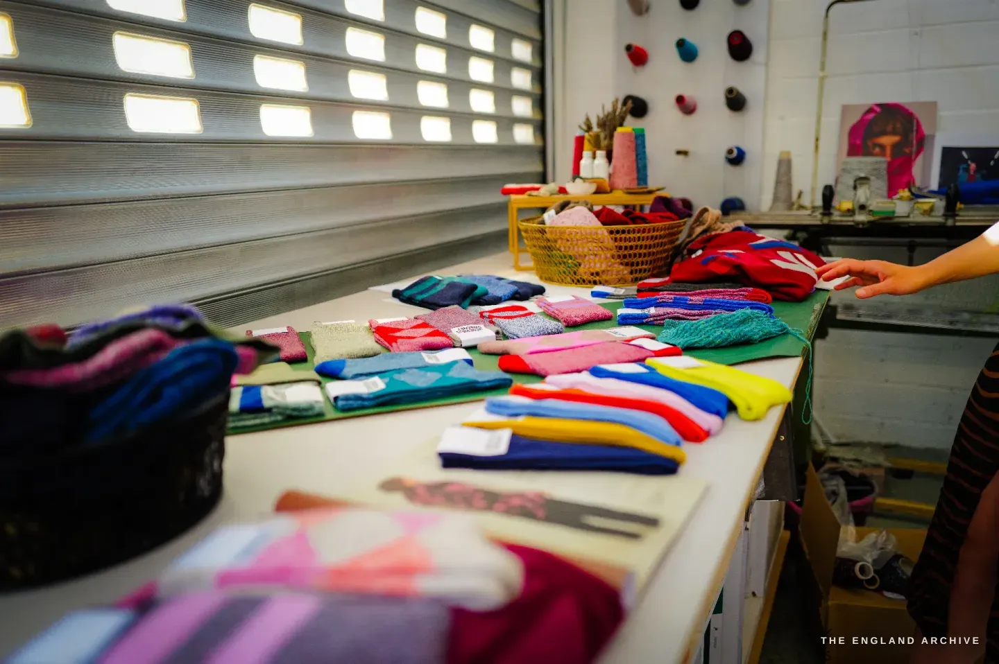 A long table laid with folded knitwear in many colours, a basket of yarn and a wall rack of cones behind, a hand reaching in from the right.