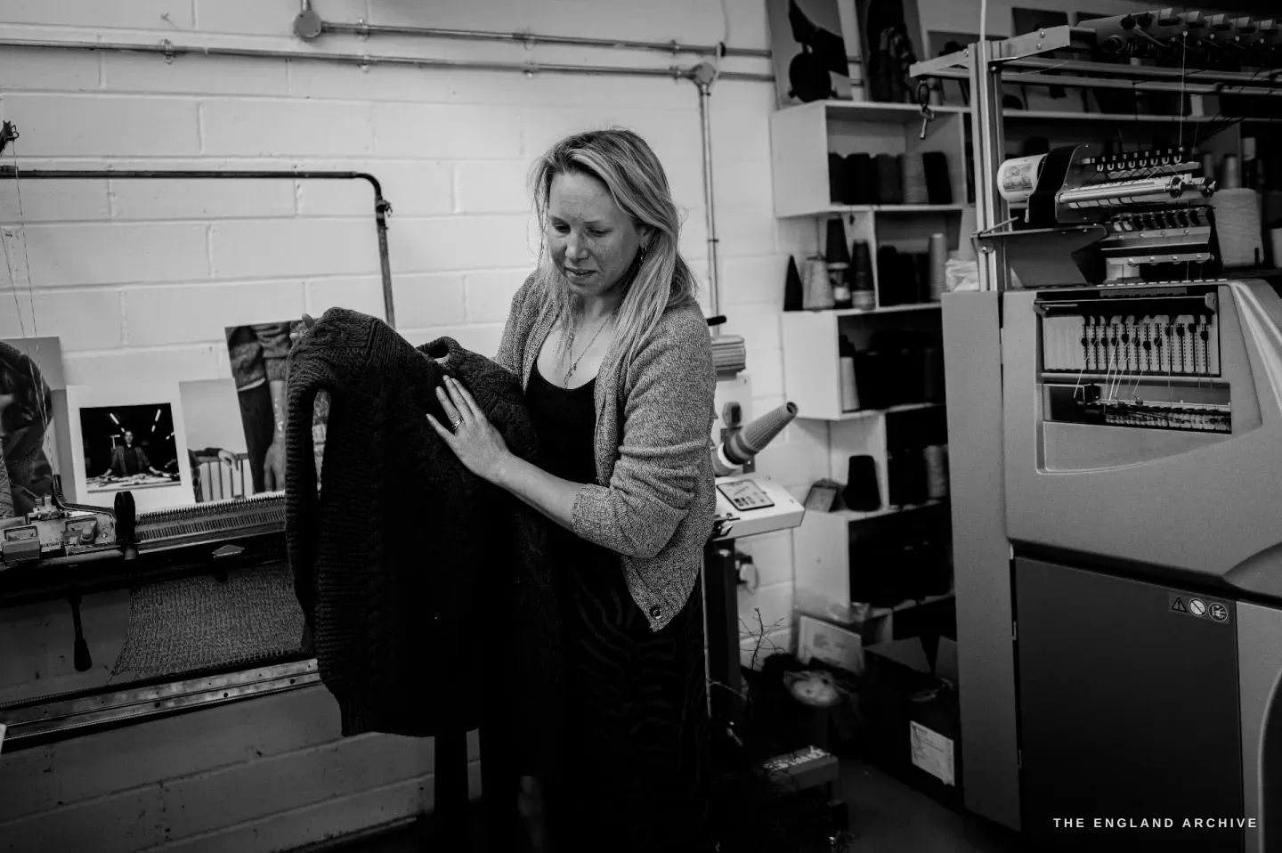 Genevieve holding a dark knitted jumper up to look at it closely, the large knitting machine to her right.