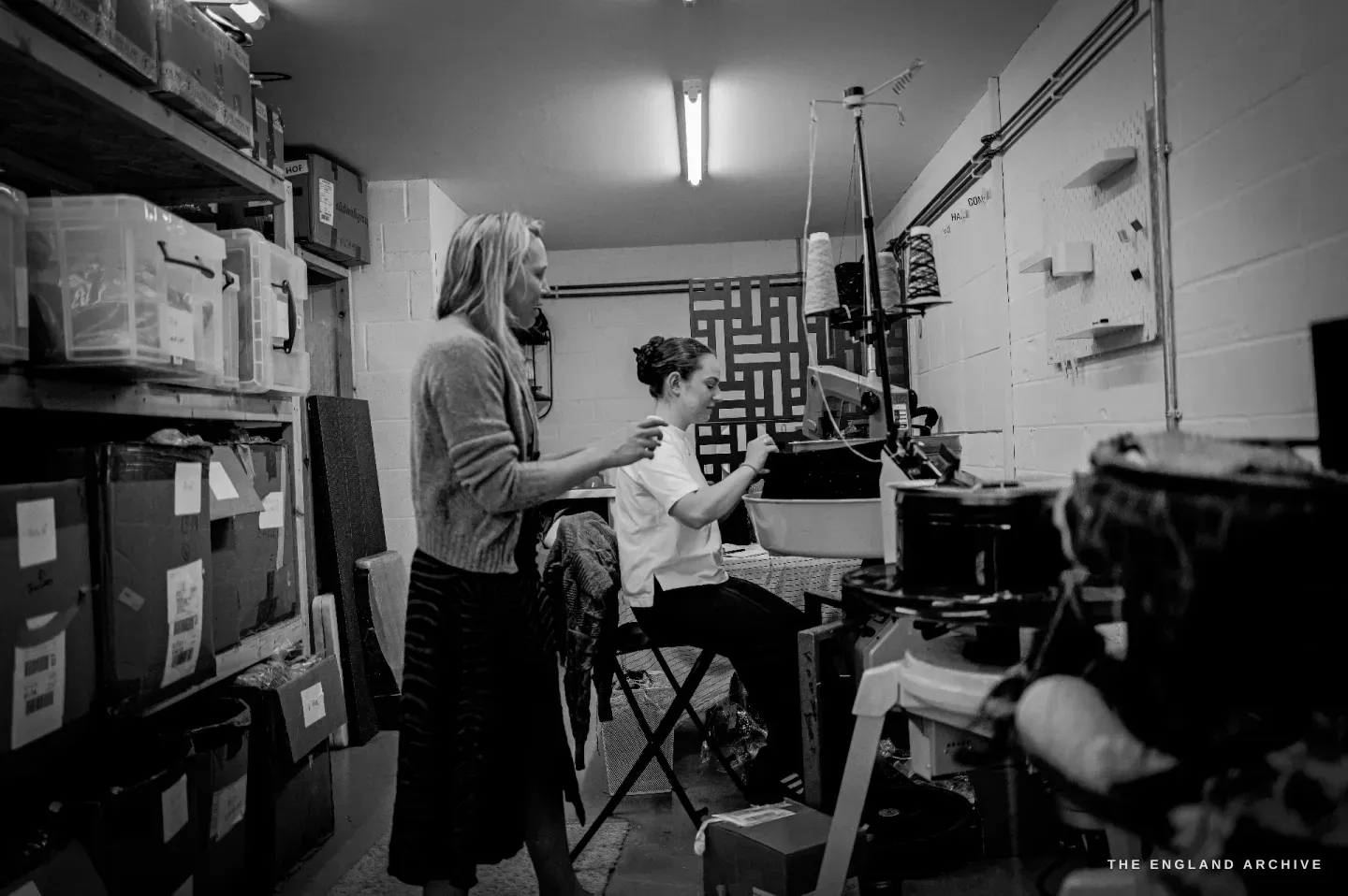 A wide view of the studio: Genevieve standing at left, Poppy seated at a machine at right, shelves of storage boxes along the wall.