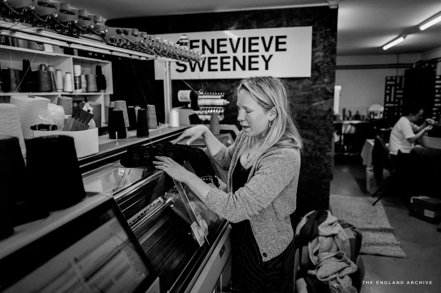 Genevieve working at a flat-knitting machine, hands on the carriage, the GENEVIEVE SWEENEY sign and racked cones of yarn above her.