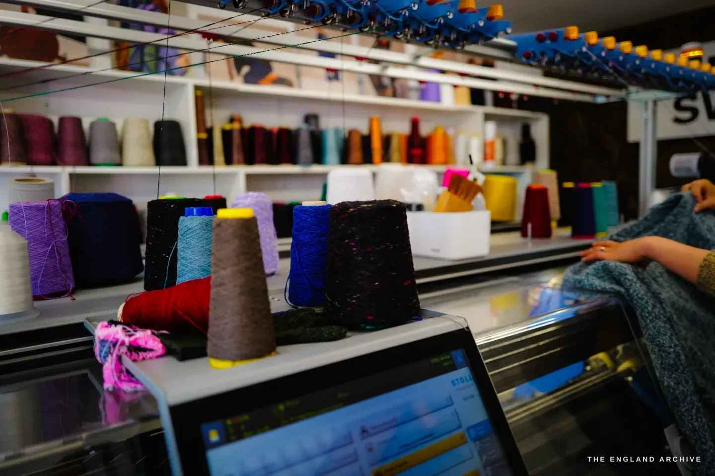Cones of coloured yarn - red, purple, teal, yellow - standing on and above the knitting machine, threads running down into the bed, the control screen lit in the foreground.