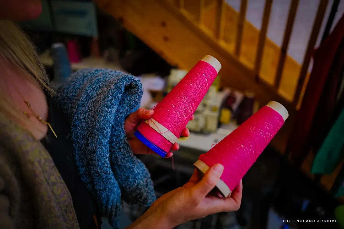 Genevieve holding up two cones of bright pink yarn, a grey knitted swatch over her shoulder.