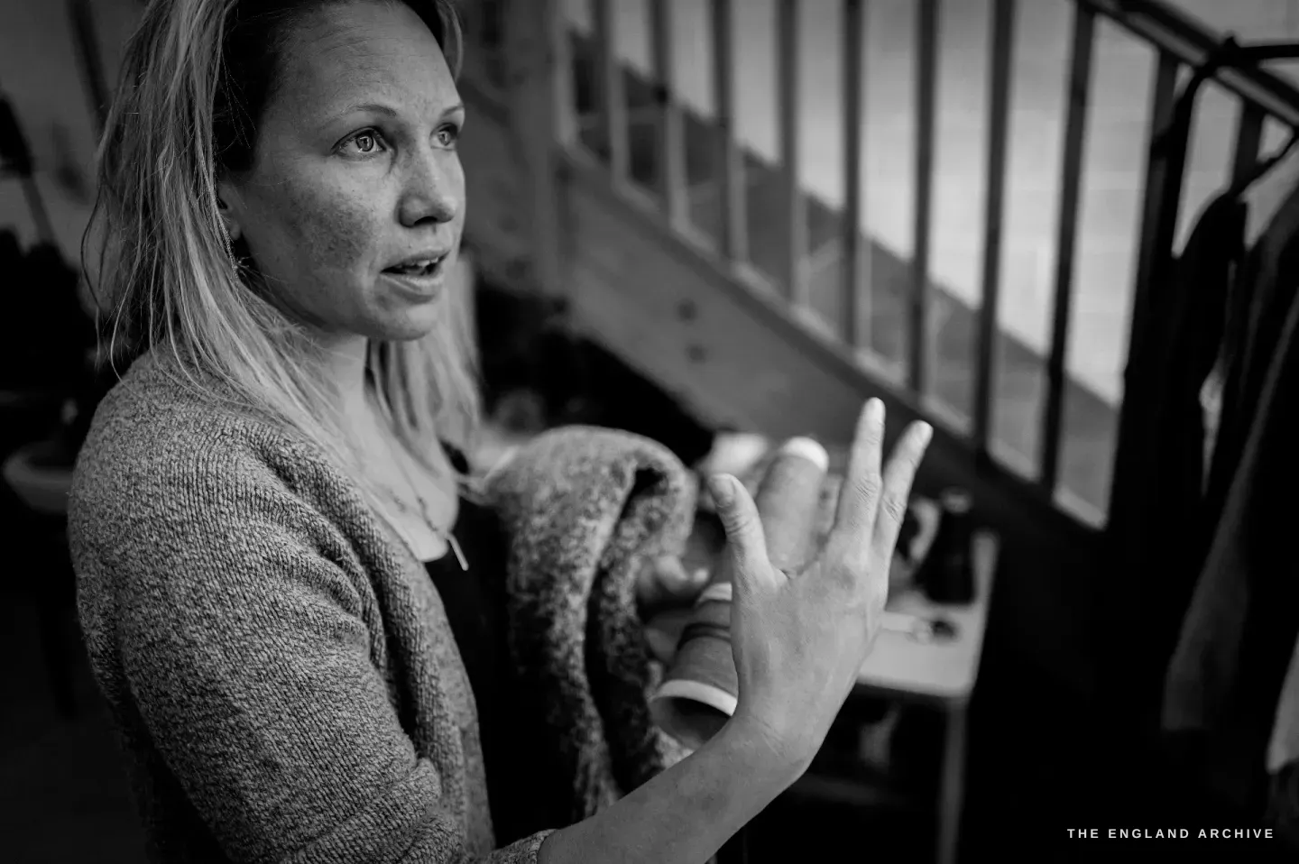 A close portrait of Genevieve talking, one hand raised, a wooden staircase behind her.