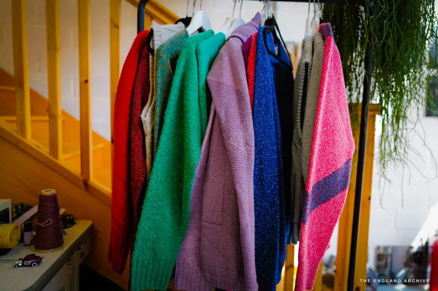 A rail of knitted jumpers in red, cream, green, lilac, blue and rose, hung against a yellow wooden staircase, a trailing plant to one side.