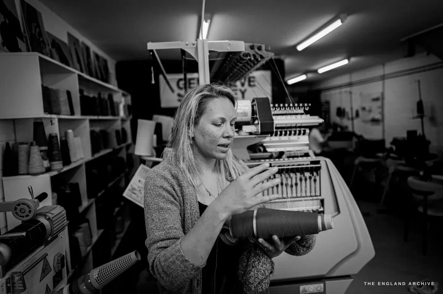 Genevieve at a cone-winding machine, winding yarn onto a cone, shelves of yarn behind her.