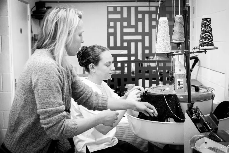 Genevieve leaning in beside Poppy, who sits at a circular sock-knitting machine threading yarn from a cone overhead.