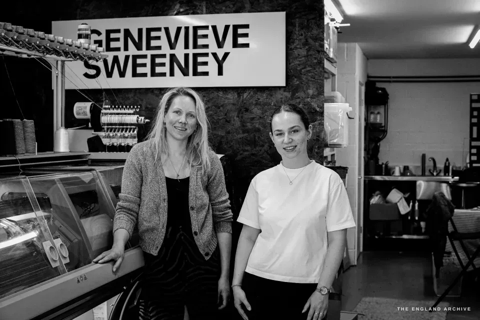 Genevieve and Poppy standing together beside the large knitting machine, the GENEVIEVE SWEENEY sign on the wall behind them.