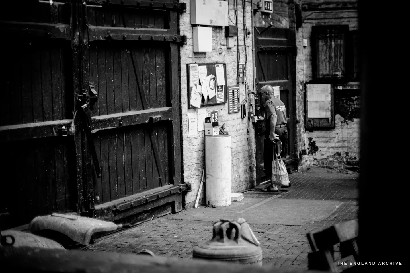 A figure with a bag standing in a doorway across a cobbled foundry yard, seen through a gap between dark wooden doors, old brick all around.