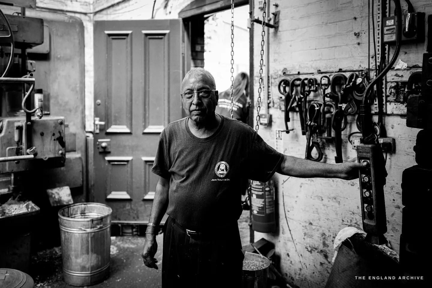 Giridhar standing in the doorway of the tuning shop, one hand resting on the machine, a green panelled door and hanging headstocks behind him.