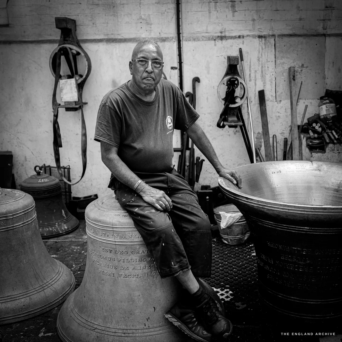Giridhar sitting on the rim of an inscribed bell, one hand flat on the bright turned lip of another bell beside him, headstocks and tools hanging on the whitewashed wall behind.