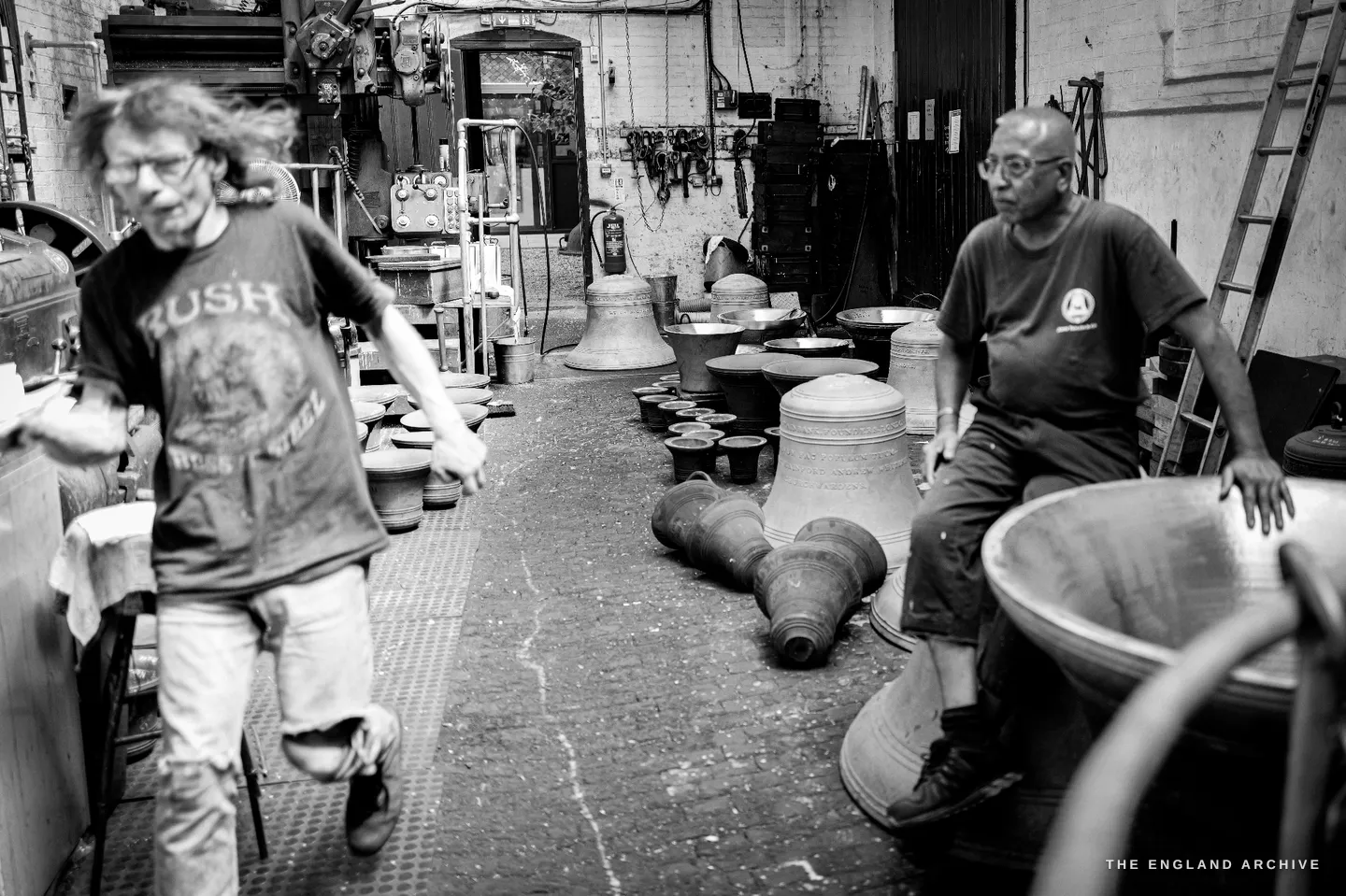 A long-haired colleague in a Rush band t-shirt striding past in motion blur while Giridhar sits among rows of bells on the right.