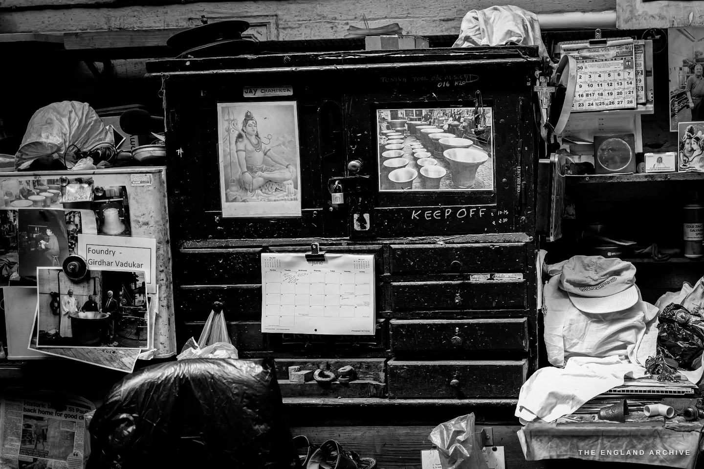 Giridhar’s workstation cabinet: a taped label reading ‘Foundry / Giridhar Vadukar’, a small printed image of a Hindu deity pinned beside photographs of bells, and a flat cap resting on the shelf.