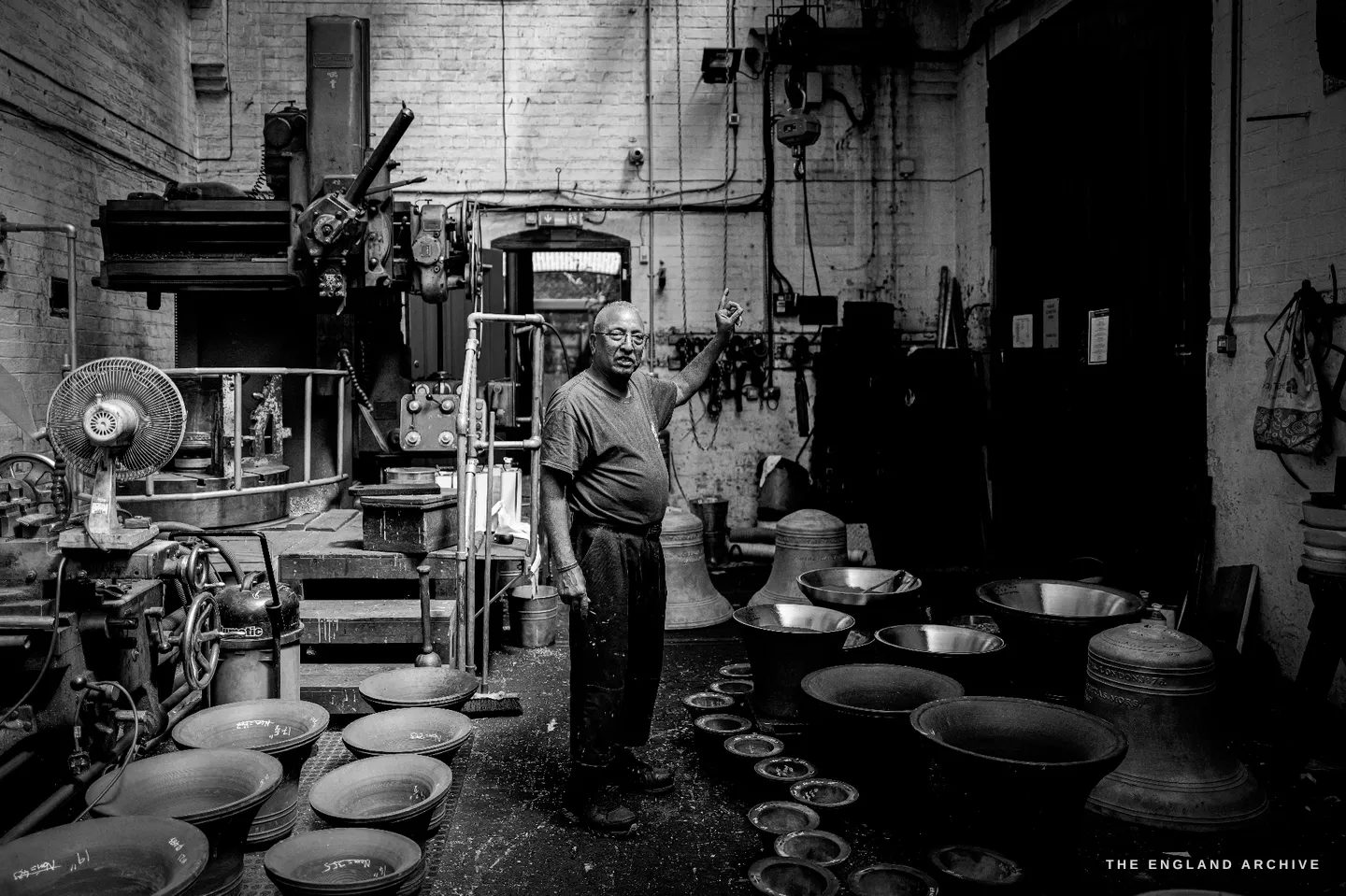 Wide view of the tuning shop with Giridhar standing at its centre, one arm raised, surrounded by dozens of bells and bowls and the tuning machinery, an electric fan to one side.