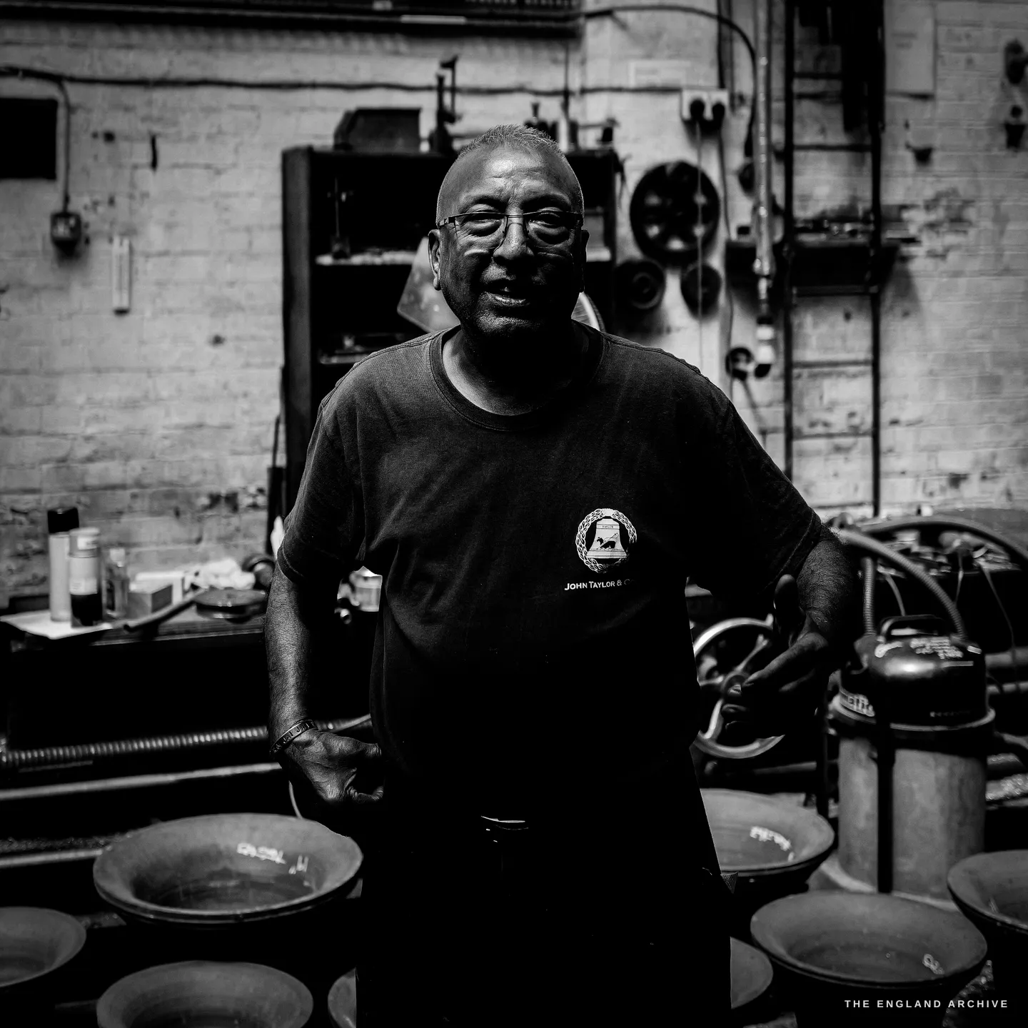 Giridhar facing the camera, one hand on his hip, the John Taylor & Co bell mark on his t-shirt, small bells ranked on the floor in front of him.