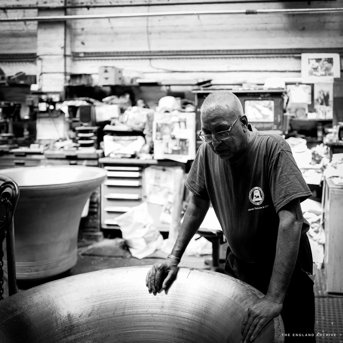 Giridhar bent over a large bell mouth-upward, both hands resting on its rim as he examines the lip, the cluttered tuning shop behind him.