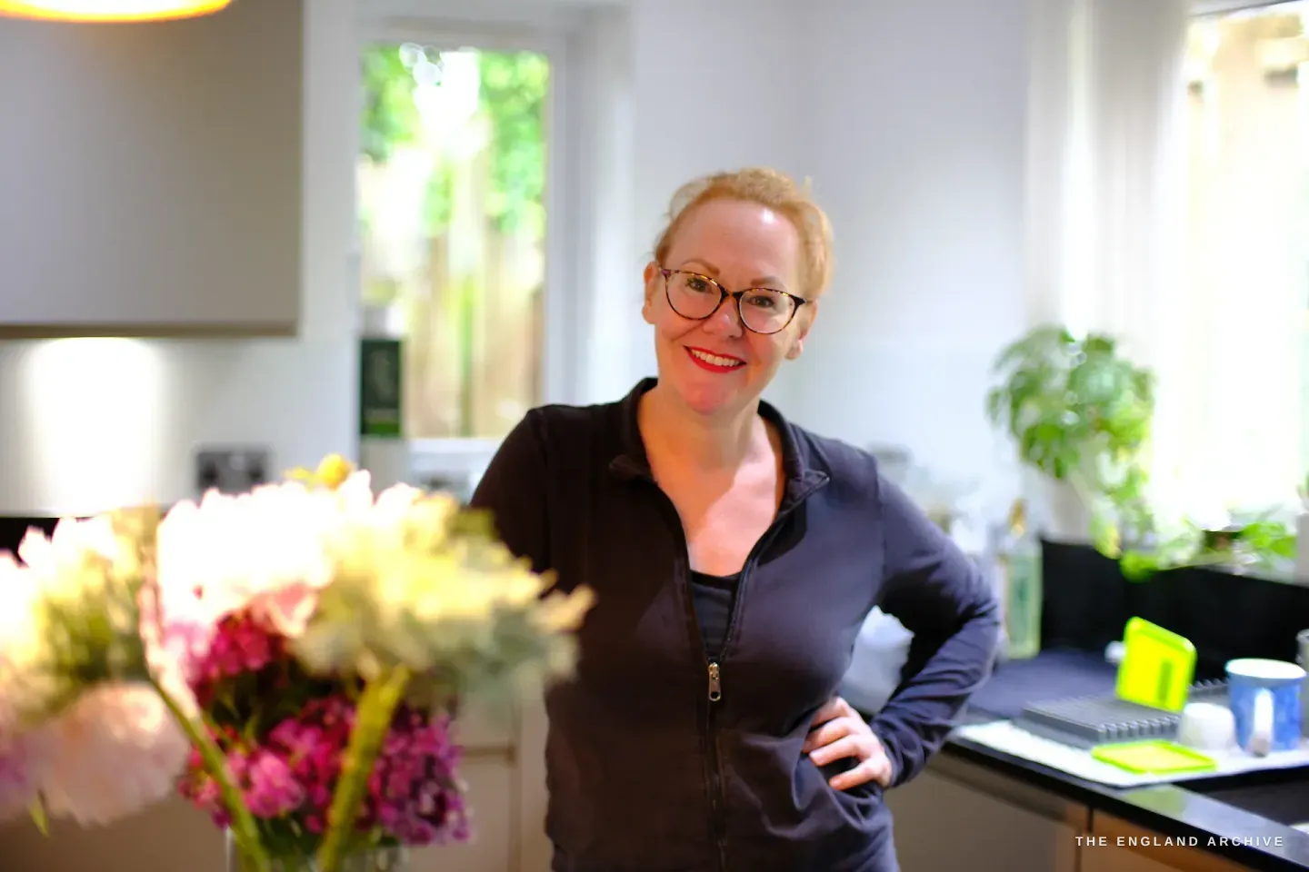 Grace Horne stands smiling in her bright kitchen, one hand on her hip, vases of pink peonies on the island in front of her and plants on the sunlit windowsill behind.