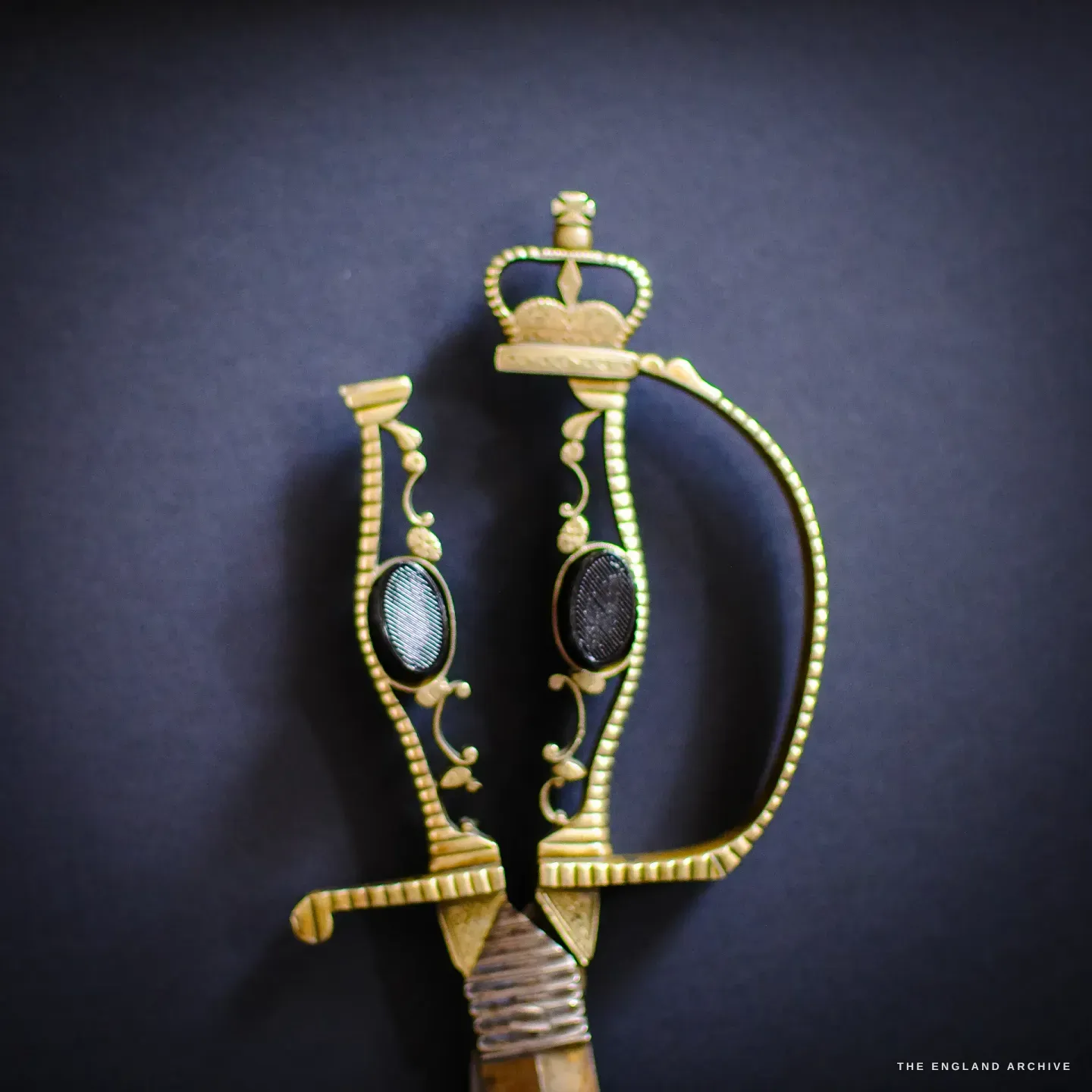 Close on the gilt basket hilt of the scissor sword - an ornate brass guard with a crown motif and oval medallions, against dark blue.