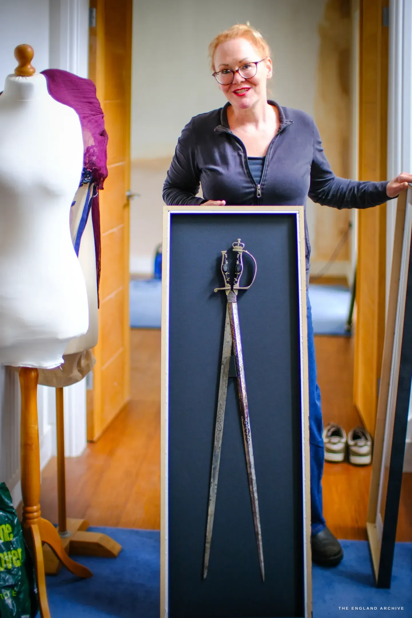 Grace Horne stands at home holding a tall framed case containing the scissor sword - a sword whose twin blades open like a pair of scissors - beside a dressmaker&rsquo;s mannequin wearing a purple corset.