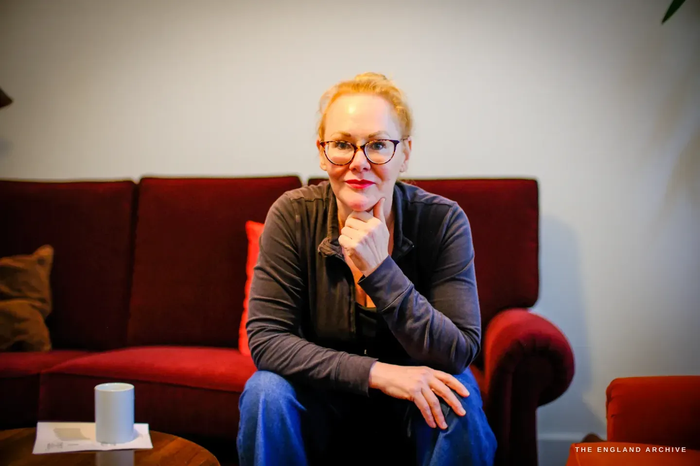 Grace Horne sits on a deep red sofa at home, hand to her chin, a mug and papers on the coffee table in front of her.