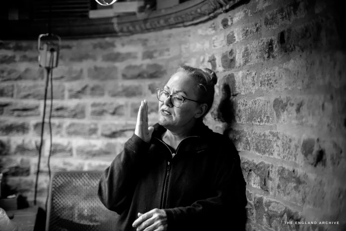 Grace Horne talks animatedly in her workshop, one hand raised mid-gesture, against the exposed stone-brick wall.