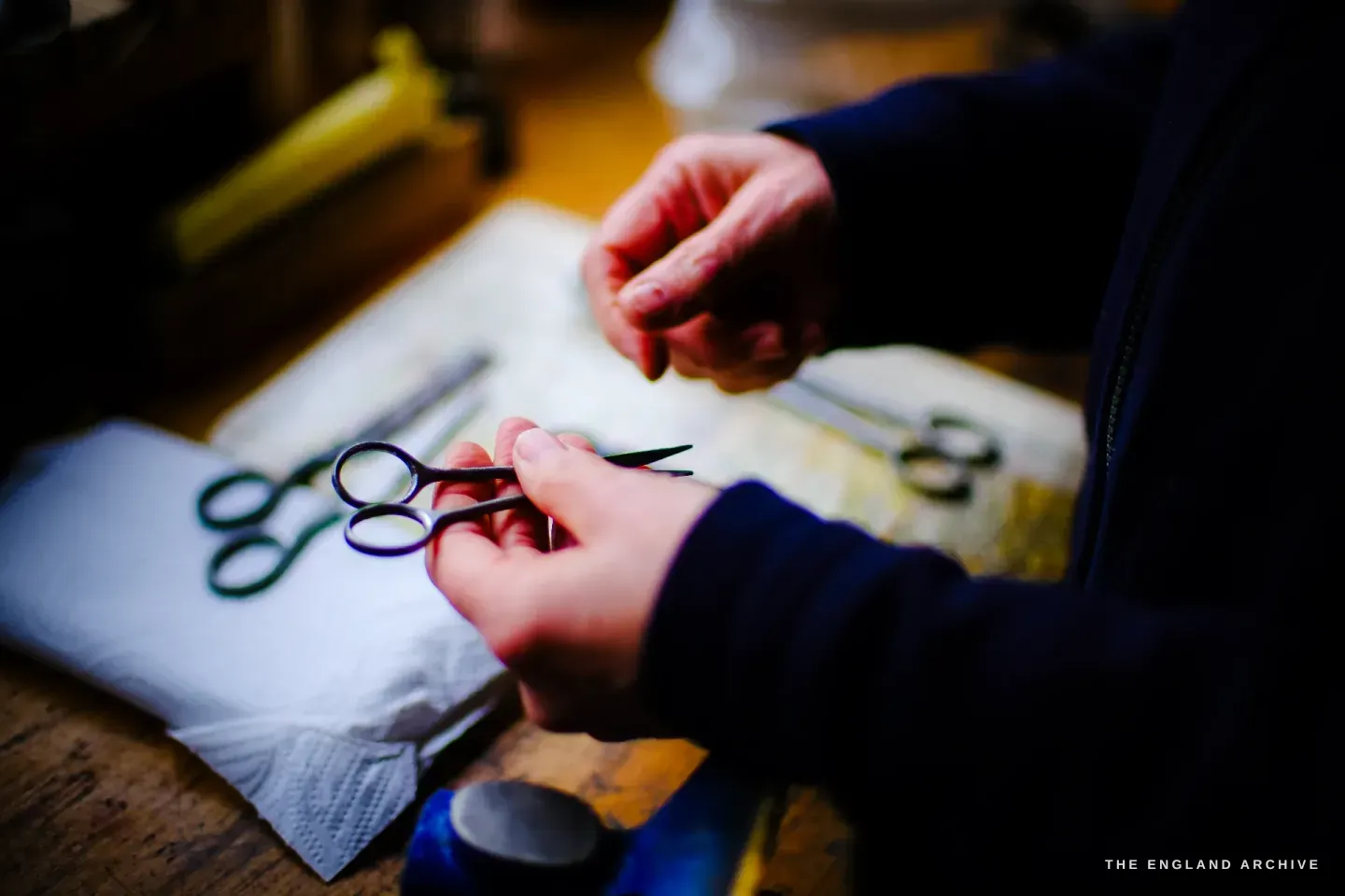 Grace Horne&rsquo;s hands hold a small pair of scissors at the bench, another small pair resting on a white cloth, in warm workshop light.