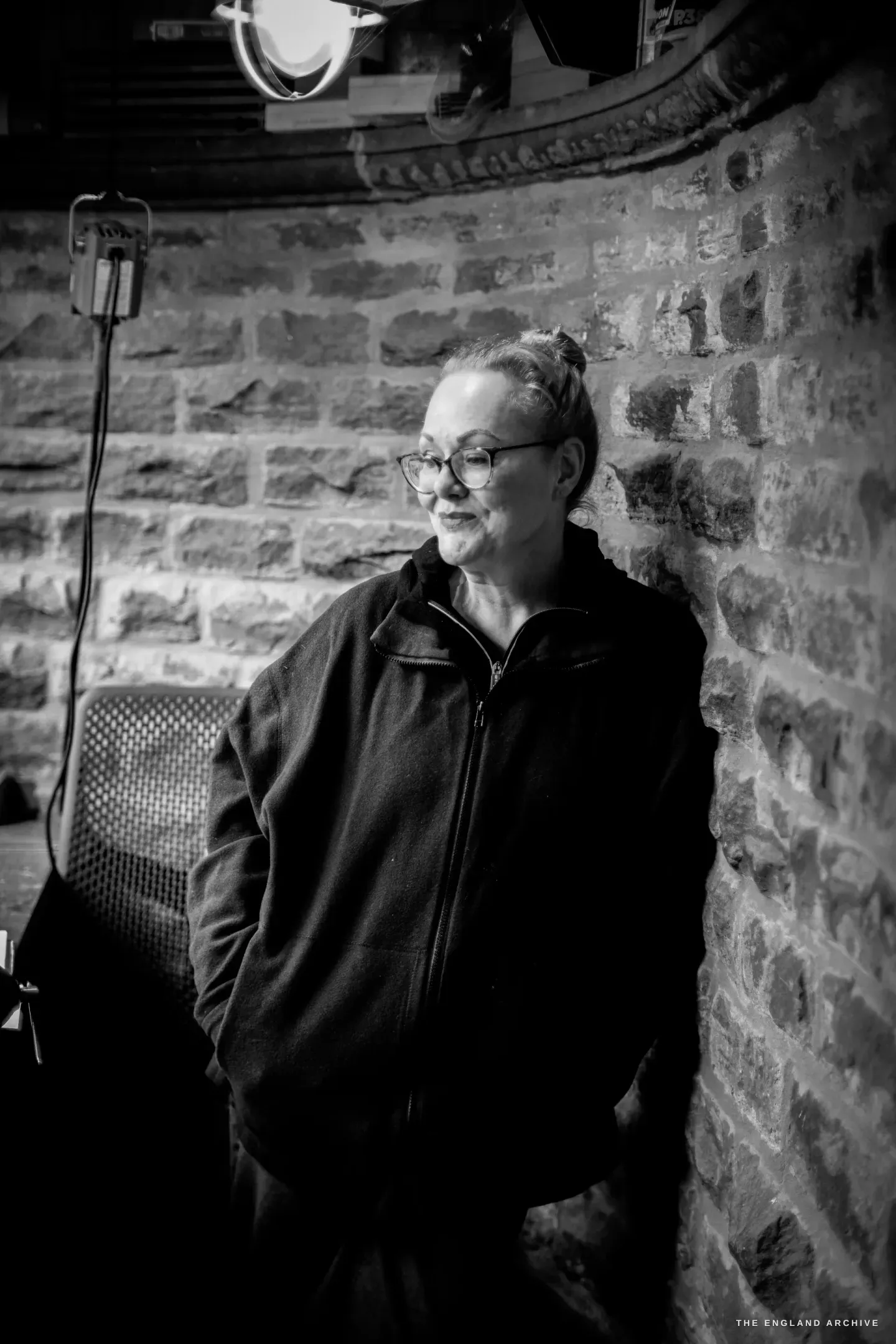 Grace Horne leans against the curved brick wall of her workshop, hands in her pockets, looking off to one side, an anglepoise lamp beside her.
