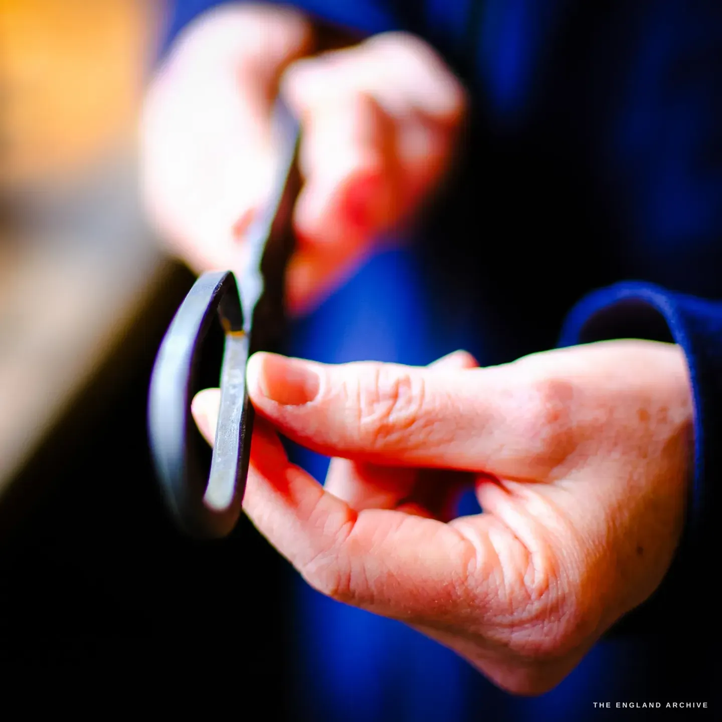 Close on Grace Horne&rsquo;s hands turning and inspecting the forged bow of a pair of scissors, her blue sleeve and the warm bench beyond.