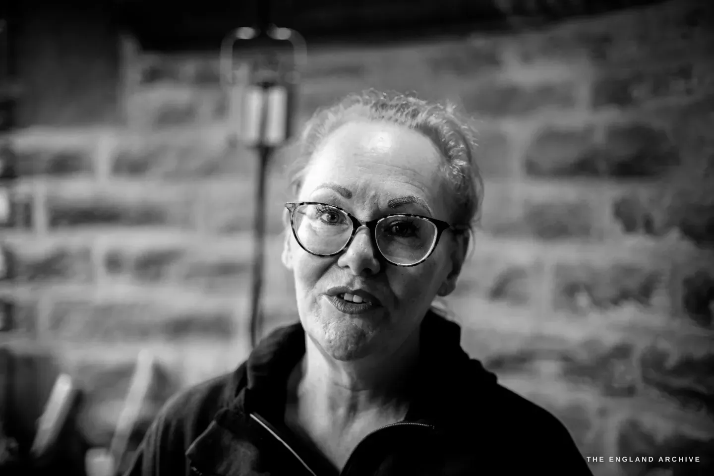 A close portrait of Grace Horne mid-speech in the workshop, glasses on, a hanging lamp and the stone wall behind.