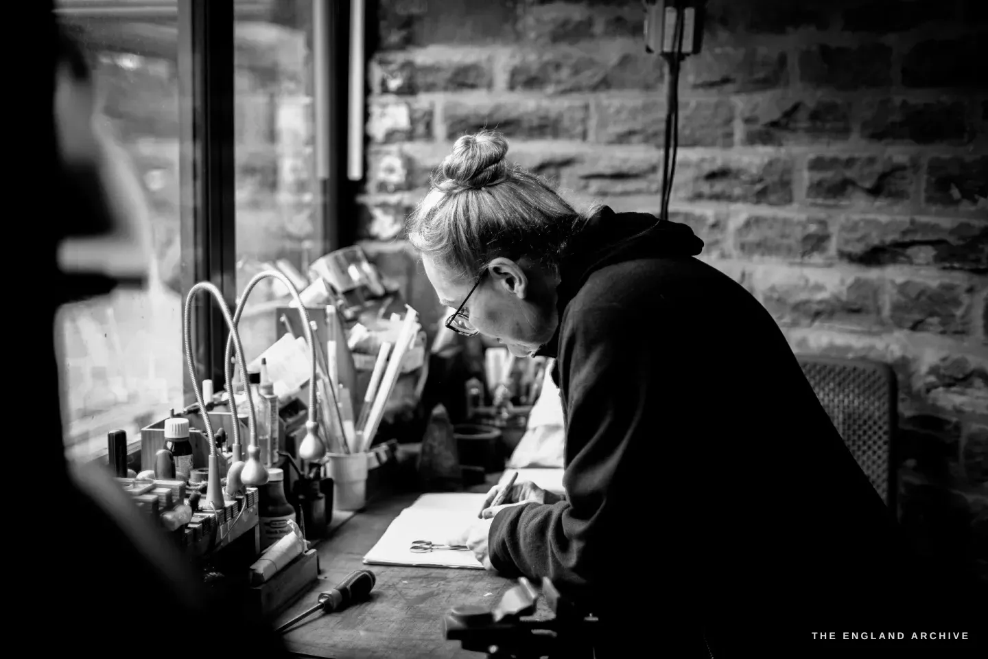 Grace Horne bends over her bench beneath the window, working, the bench lined with files and scribes in racks against the brick wall.