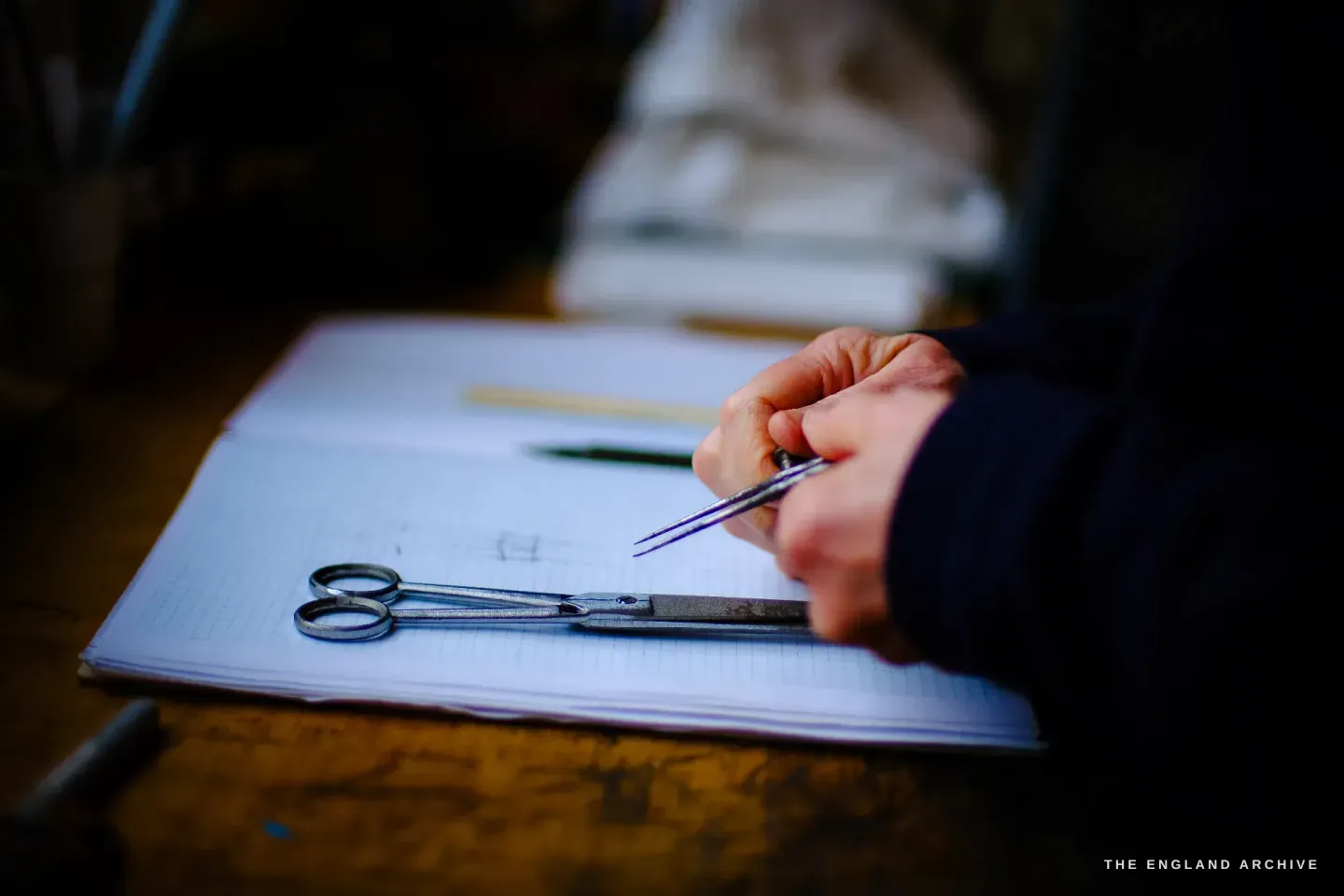Grace Horne uses a pair of dividers to step out a measured scissor design on graph paper, a finished pair lying on the sheet for reference.