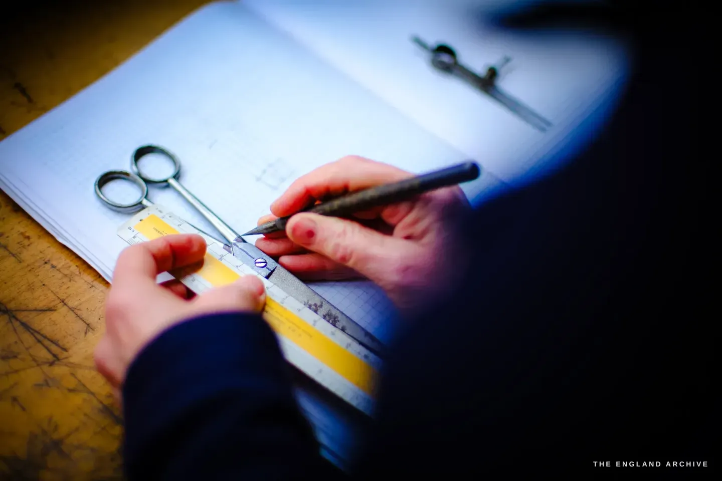 Grace Horne marks a measured drawing on graph paper with a scriber against a steel rule, a small pair of scissors and dividers beside her hands.