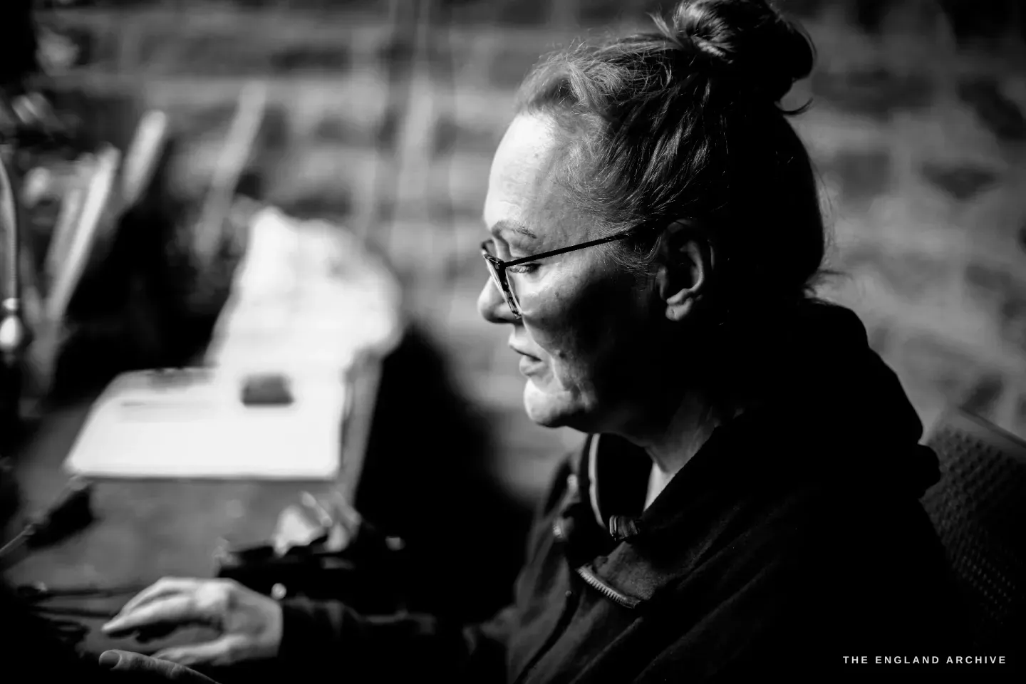 A side-lit profile of Grace Horne working intently at her bench, hand at the work on a white sheet, in deep shadow.