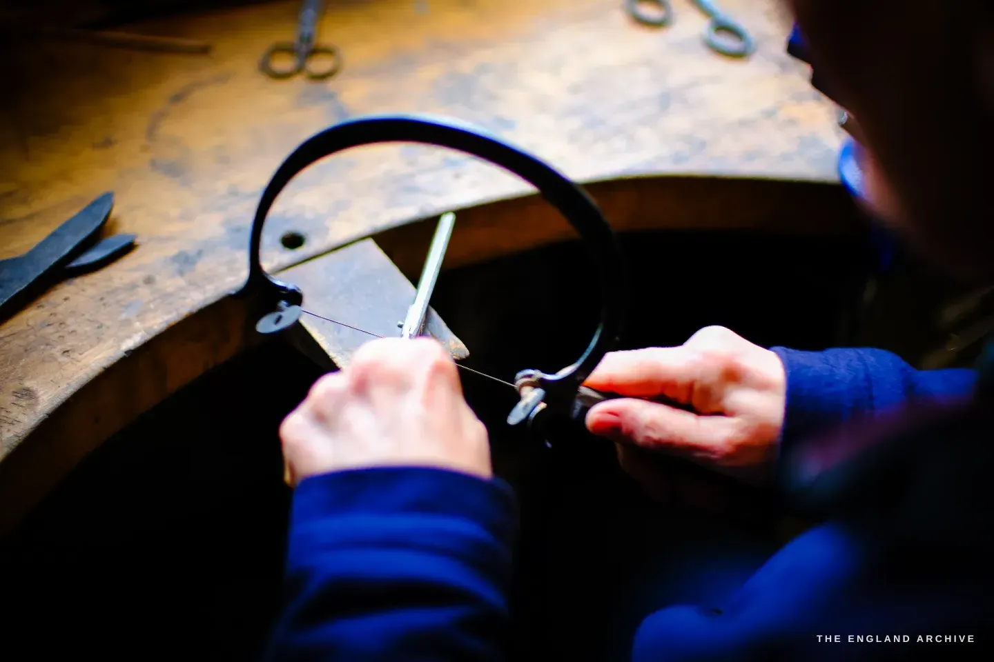 Over Grace Horne&rsquo;s shoulder, her hands use large calipers to measure across a scissor component on the bench, finished scissors beside.