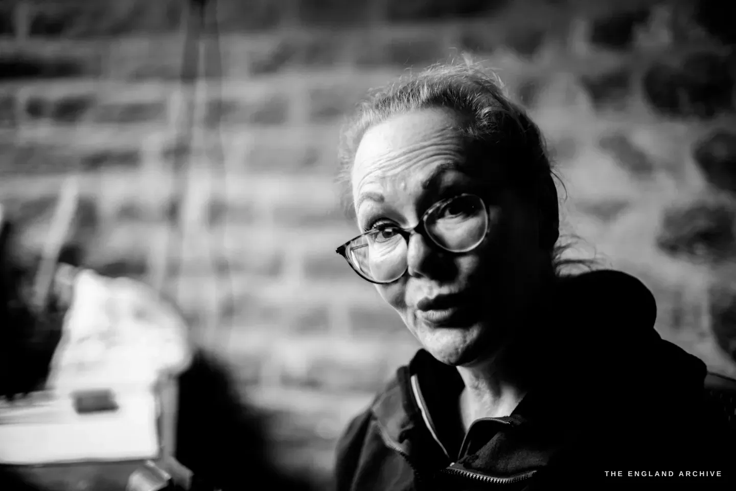 A dramatic close portrait of Grace Horne in her workshop, side-lit against the stone wall, glasses on, an intent expression.