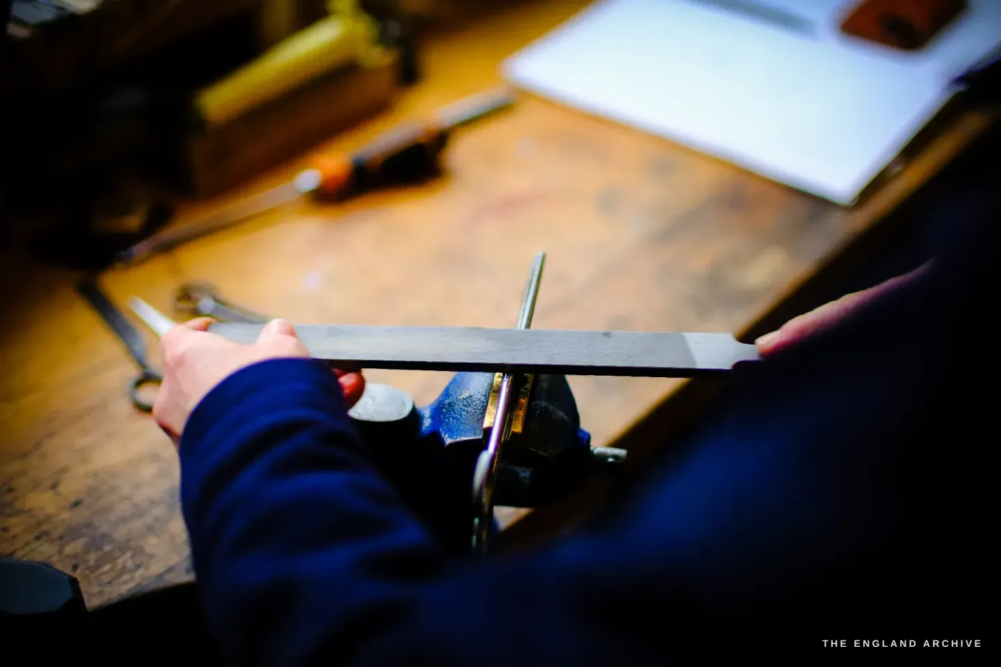 Grace Horne&rsquo;s hands file the blade of a pair of scissors held in a bench vice, tools and scissors on the warm wooden bench around her.