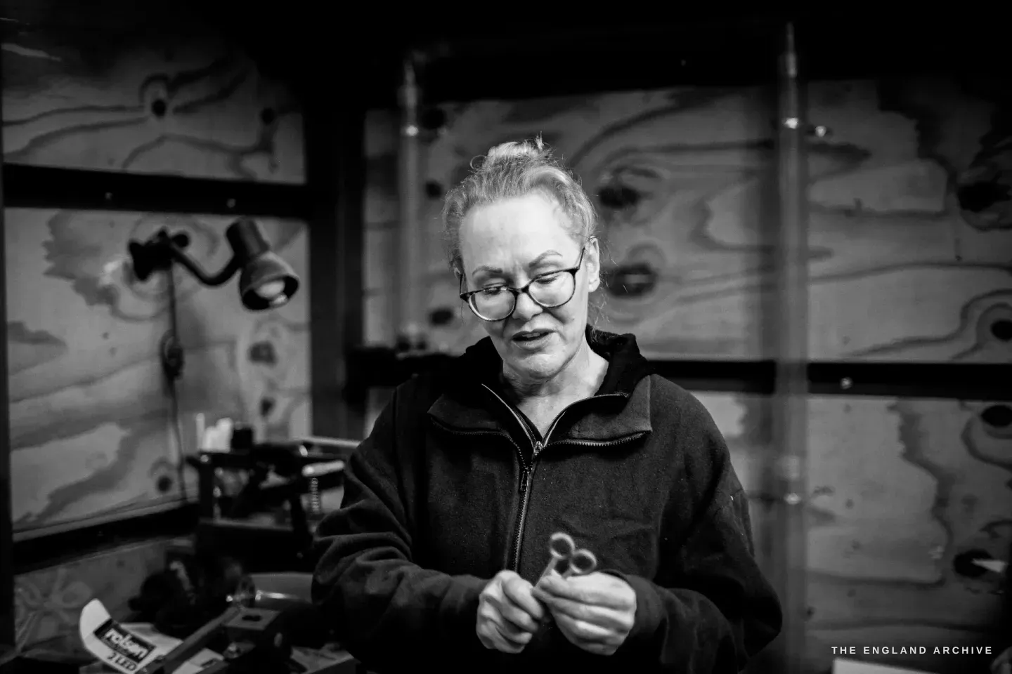 Grace Horne stands in the workshop holding a small pair of scissors, looking down at them, a plywood-lined wall behind.