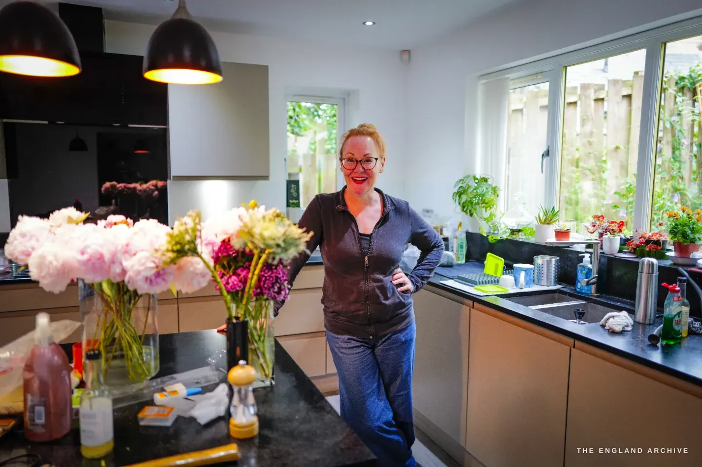 Grace Horne stands smiling in her bright kitchen, hand on hip, vases of pink peonies on the island and flowers and plants along the window.