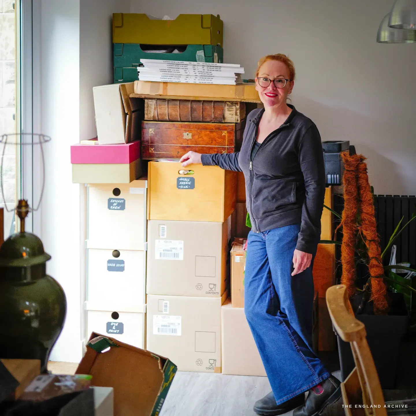 Grace Horne stands at home beside stacked, labelled archive boxes, old leather books and a wooden chest - her collection of scissor research.